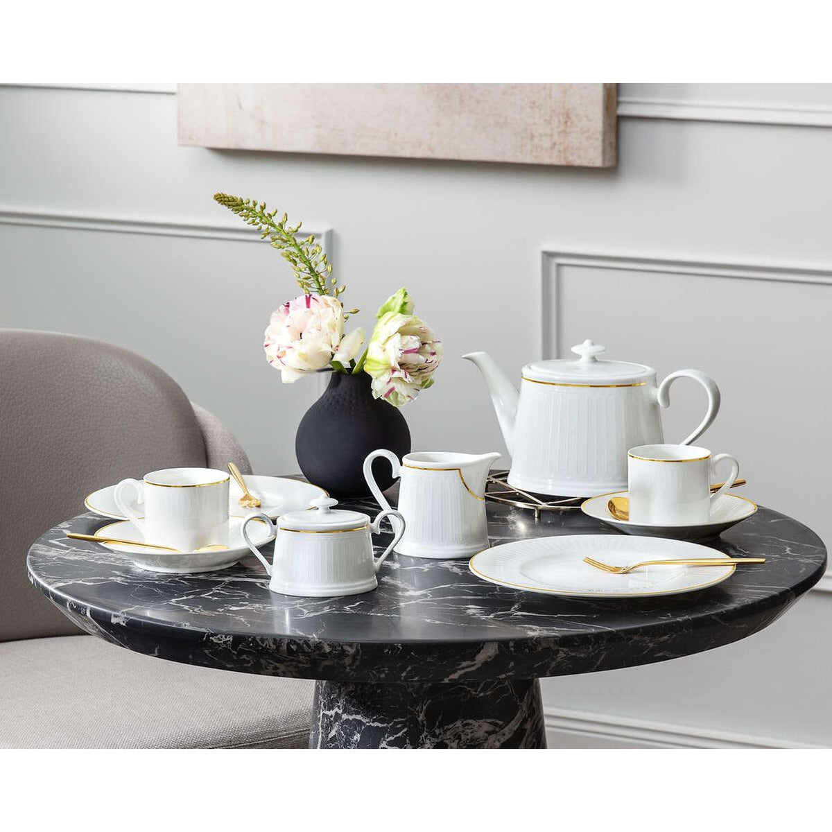 A styled tea set arranged on a round black marble table in a dining room setting. The set includes a white teapot with gold trim, multiple white teacups and saucers with gold trim, a white creamer, white plates, and gold spoons.