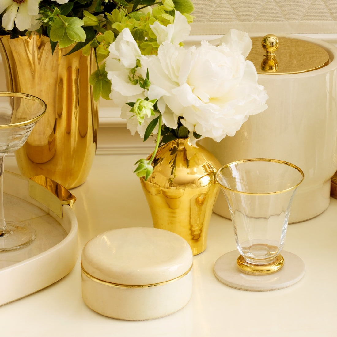 Decorative setting with gold vases, white flowers, and glassware on a light surface.