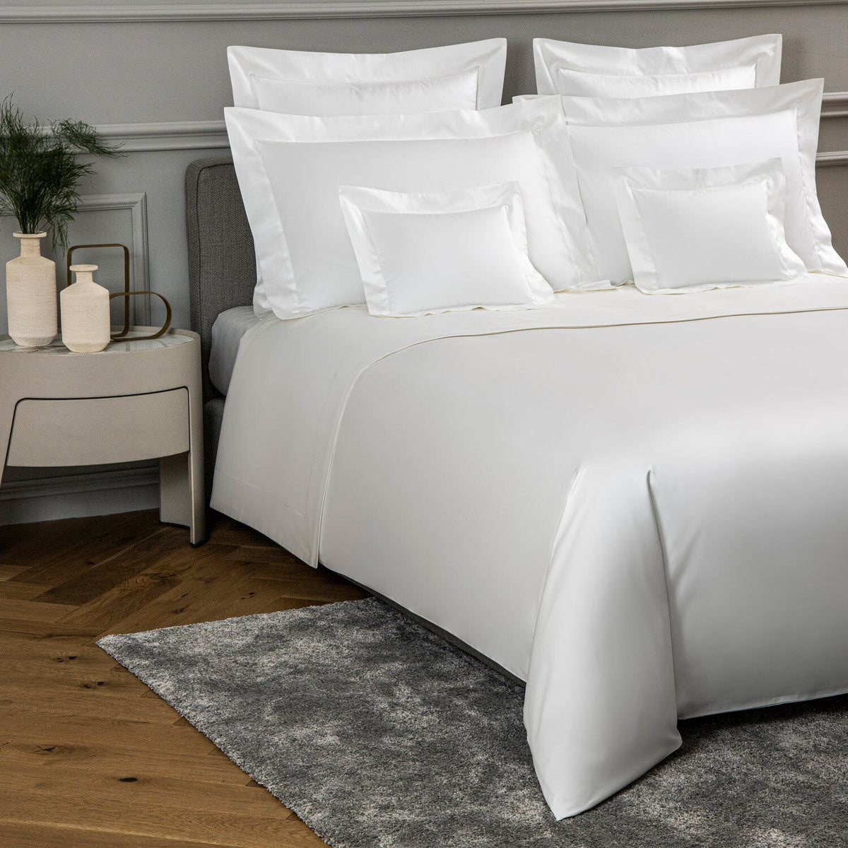 A neatly made bed with crisp white bedding, including multiple pillows and a white duvet, styled in a modern bedroom with a gray headboard, wooden floor, gray rug, and a light gray nightstand with decorative vases.