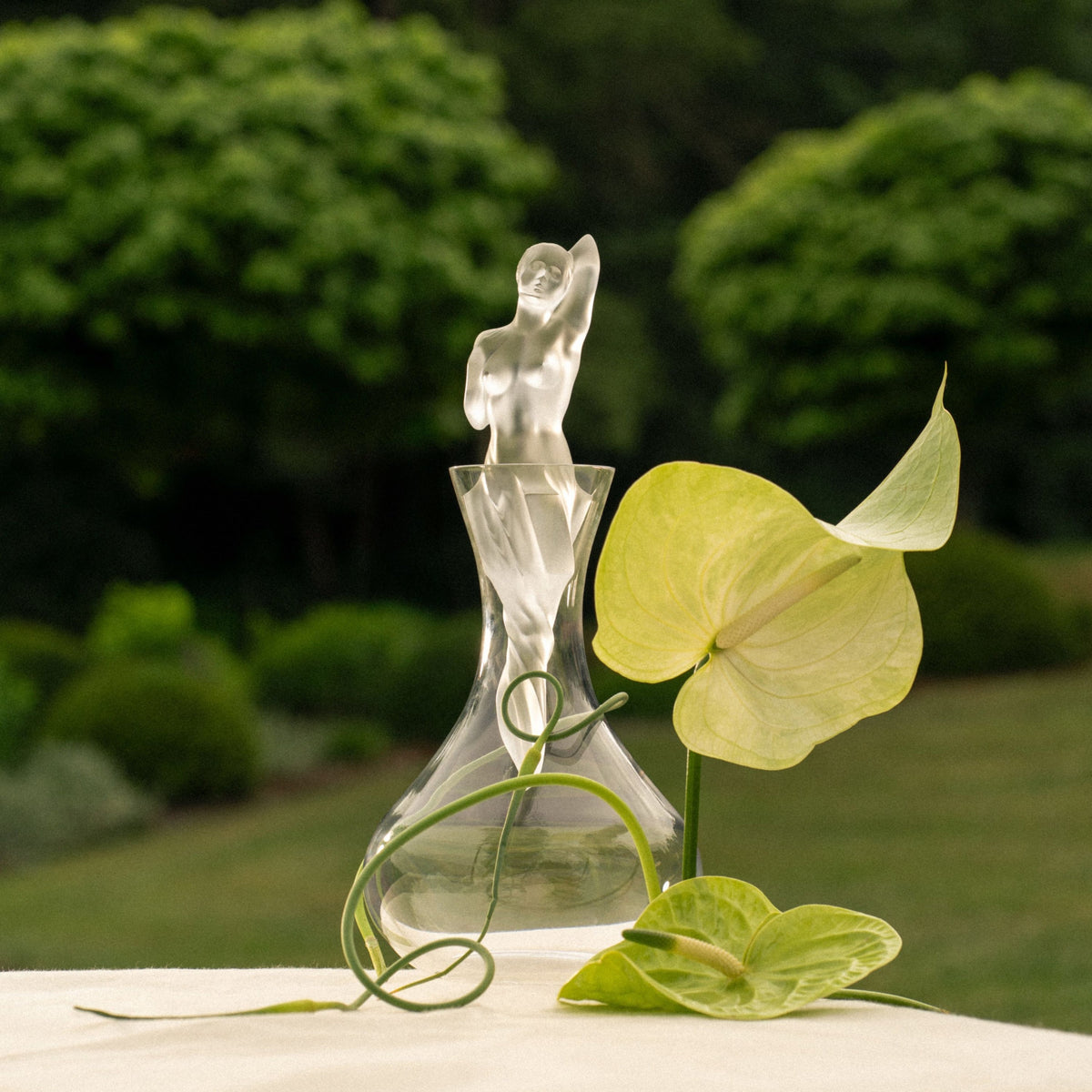 Clear glass sculpture of a woman with a plant on a table outdoors