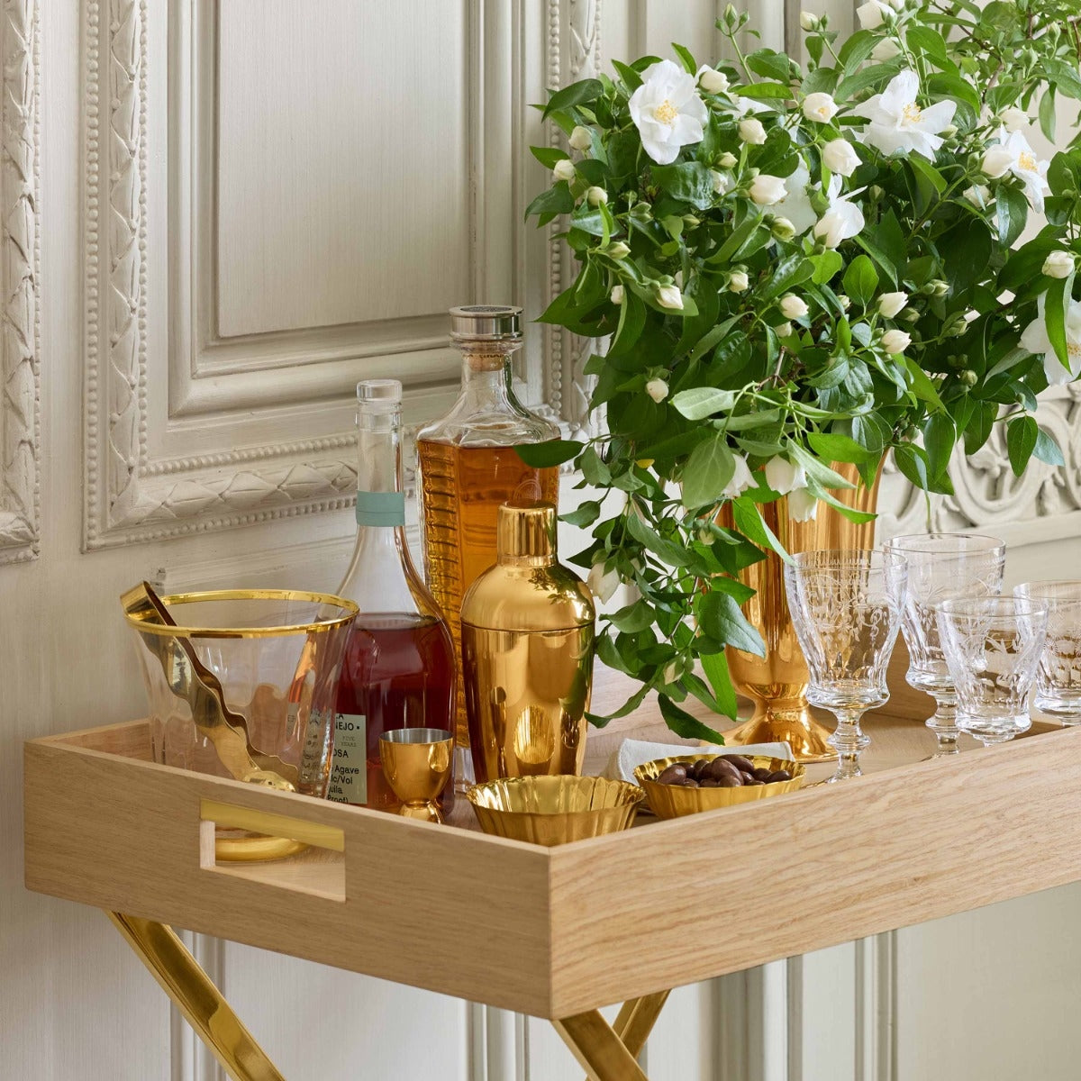 A light oak wood bar cart with gold-tone legs and handles, styled with gold and clear barware, a bottle of amber liquor, and a large green floral arrangement against an ornate cream wall.