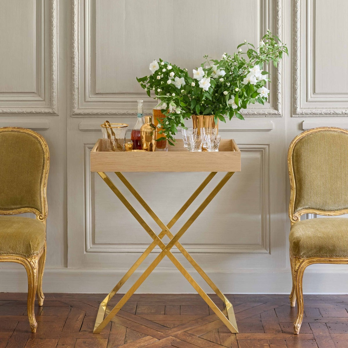 A small wooden serving tray table with a gold X-frame base, styled in a classic room with two gold chairs and a floral arrangement on top.