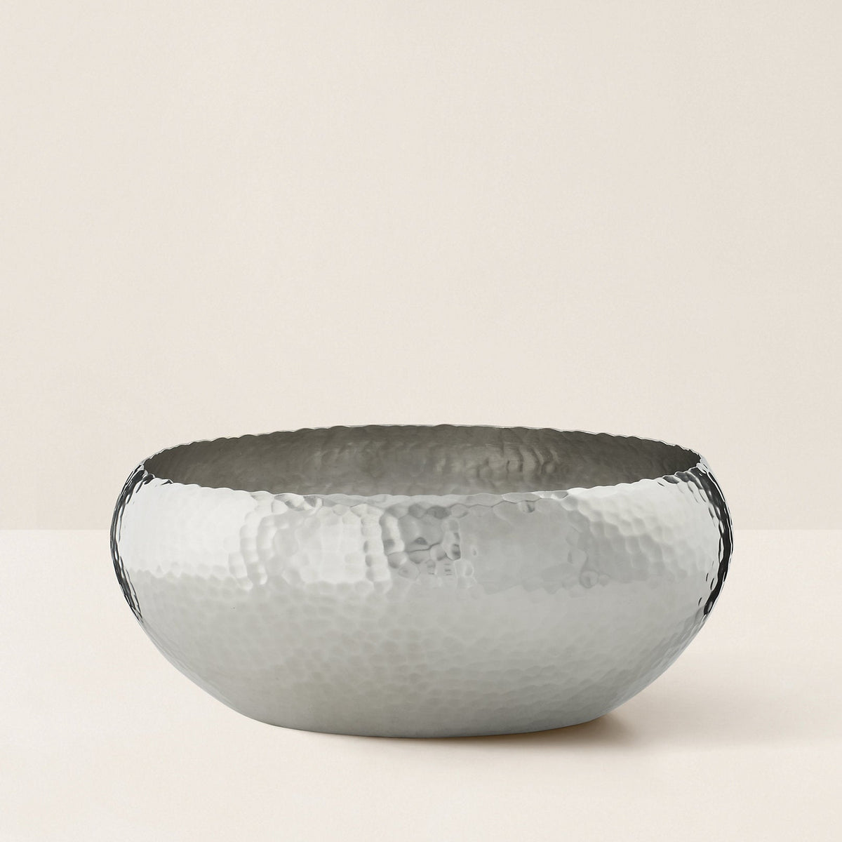 A hammered metal bowl with a textured, dimpled surface and darkened edges, photographed on a clean, solid, warm off-white background.
