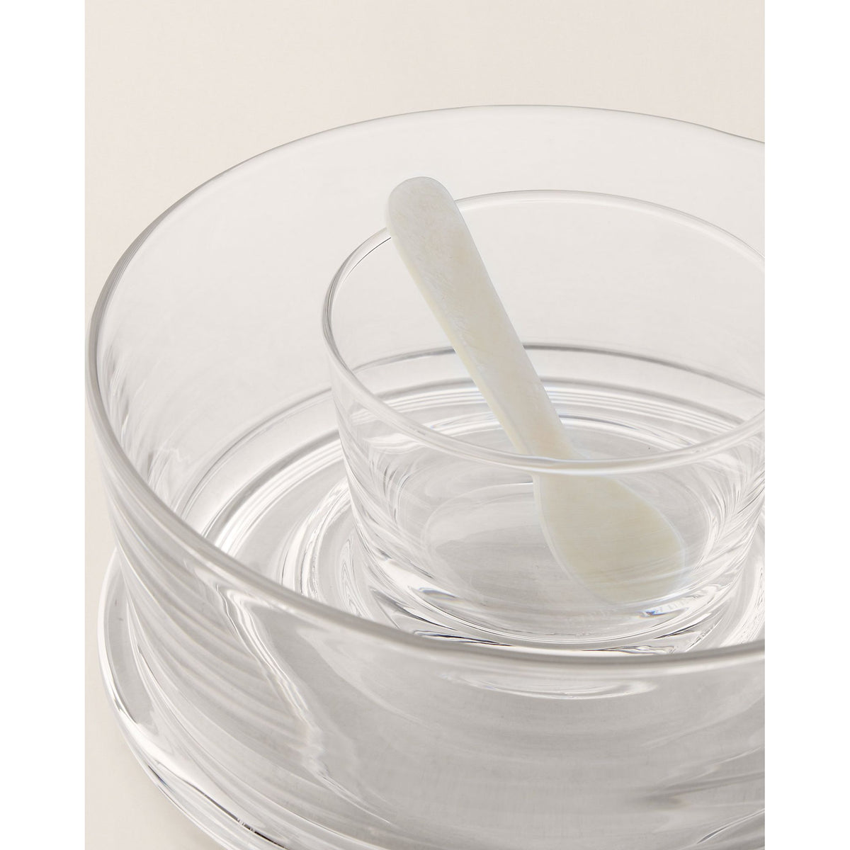 Clear glass bowl with a white spoon inside on a light beige background