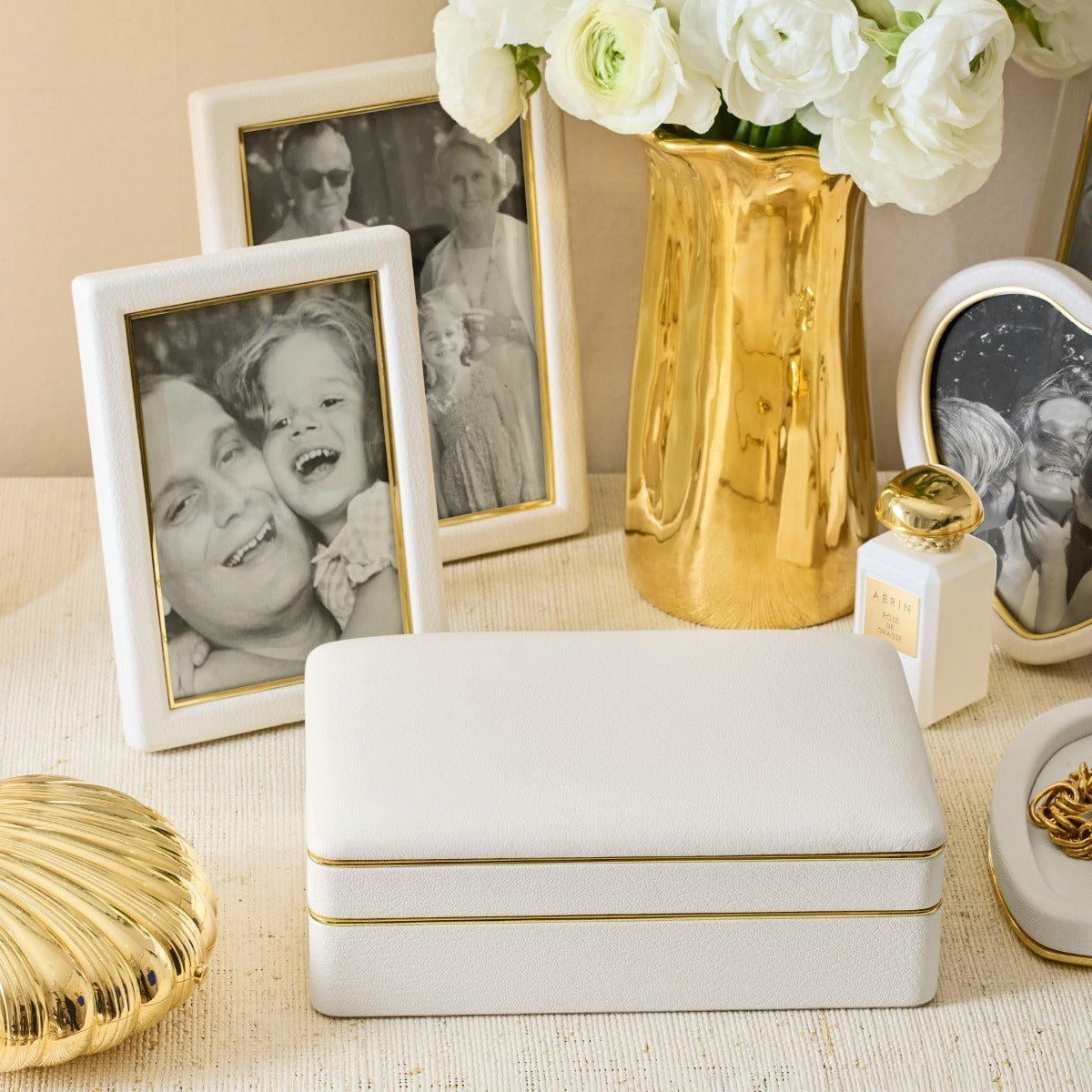 A rectangular white jewelry box with thin gold trim sits on a light beige surface, styled with black-and-white photo frames, a gold vase of white flowers, a small gold perfume bottle, and a gold shell-shaped decor piece.