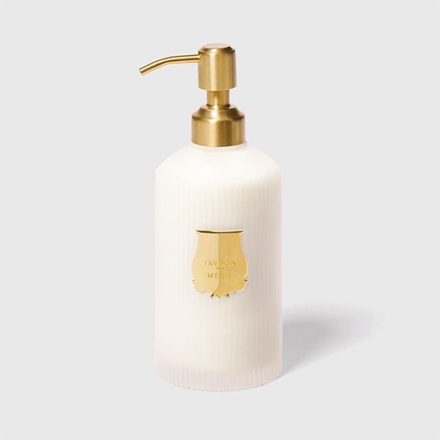 A frosted white ribbed pump bottle with a polished gold pump dispenser and a matching gold shield-shaped logo plaque on the front, set against a light gray background.