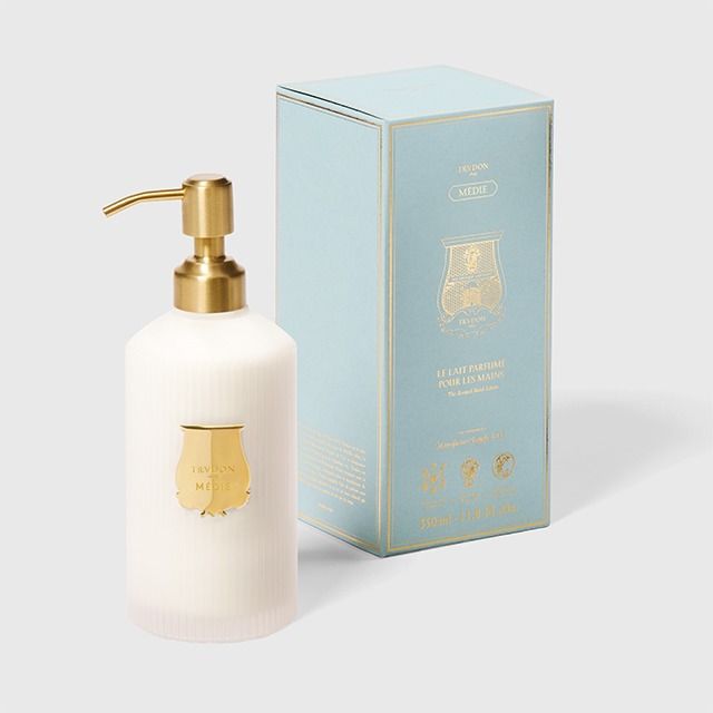A frosted white pump bottle with a gold pump and gold shield emblem, shown next to its light blue and gold branded box on a light gray background.