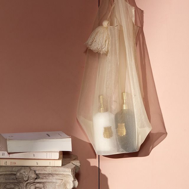 A sheer cream mesh tote bag with a tassel hanging on a pink wall, holding two pump bottles, with stacked books on a stone surface below.