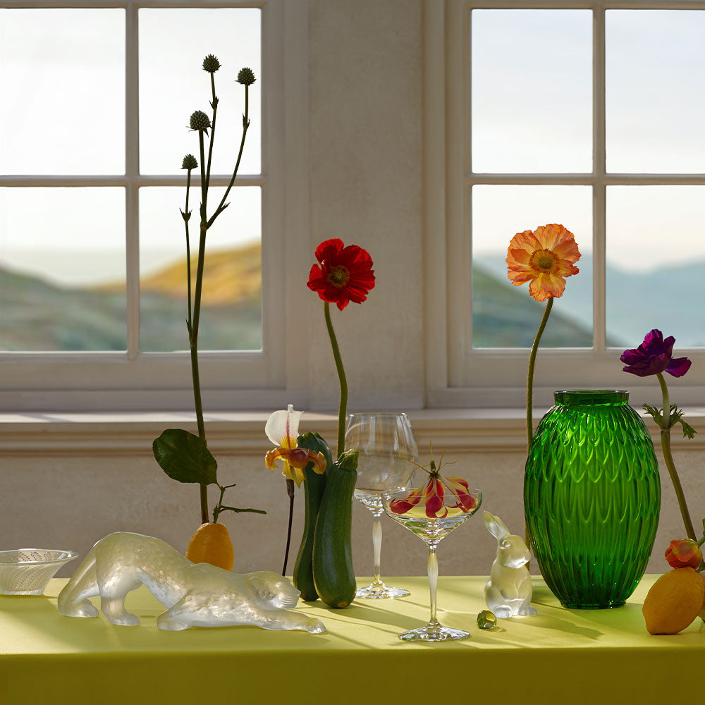Decorative table setting with flowers, vases, and glasses in a room with large windows.